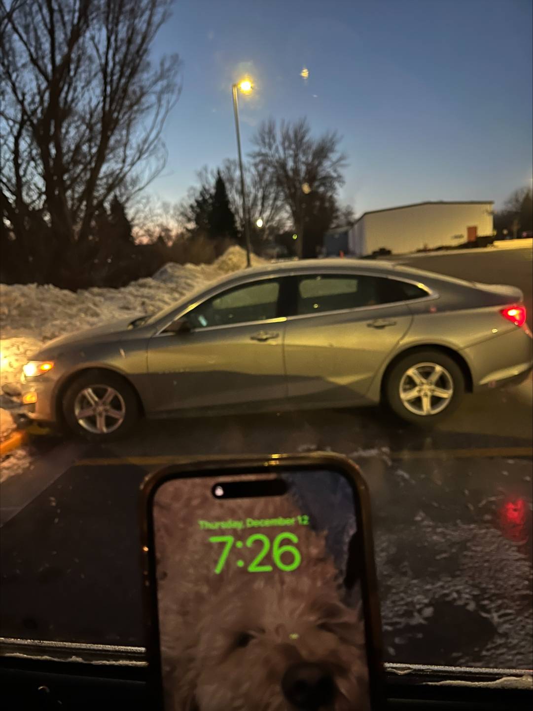 In the foreground is Erin's phone displaying the time, 7:26 a.m. In the background is the state car parked in a parking lot. The air looks chilly and there are piles of snow on the perimeter of the parking lot.